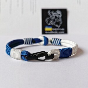 May include: A blue and white paracord bracelet with a black clasp. The bracelet is made with two different types of paracord, one with white stripes and one with blue stripes.