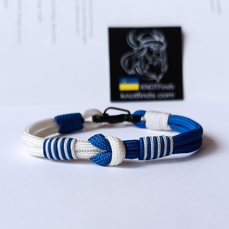 May include: A blue and white paracord bracelet with a black clasp. The bracelet has a knot in the center and is made of two strands of rope.