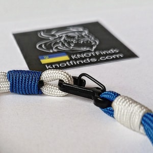 May include: A white and blue paracord bracelet with a black metal clasp. The bracelet is tied with a knot and has a Ukrainian flag charm.