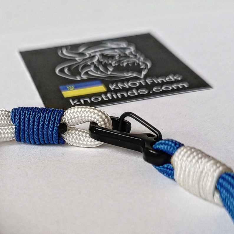 May include: A white and blue paracord bracelet with a black metal clasp. The bracelet is tied with a knot and has a Ukrainian flag charm.