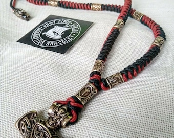 Peut inclure: Un collier en cordon tressé noir et rouge avec un pendentif Mjolnir argenté. Le pendentif présente des nœuds complexes et une rune sur le dessus. Le collier est agrémenté de perles argentées.