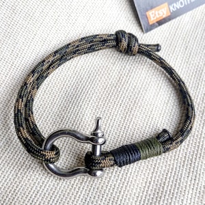 Op de afbeelding: Een groene en zwarte camouflage paracord armband met een zilveren metalen sluiting. De armband is met een knoop vastgemaakt en heeft een zwart en groen accent.