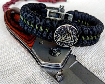 Op de afbeelding: Een zwart en groene paracord armband met een zilveren metalen bedel met een triquetra symbool. De armband ligt op een zilveren mes met een houten handvat.