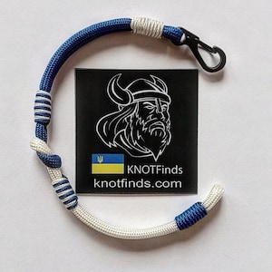 May include: A blue and white paracord bracelet with a black metal clasp. The bracelet is tied in a knot pattern. The bracelet has a small Ukrainian flag charm. The bracelet is labeled "KNOTFinds knotfinds.com".