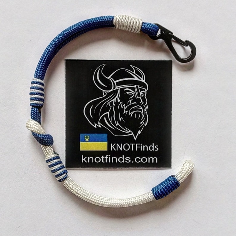 May include: A blue and white paracord bracelet with a black metal clasp. The bracelet is tied in a knot pattern. The bracelet has a small Ukrainian flag charm. The bracelet is labeled "KNOTFinds knotfinds.com".