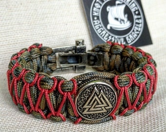 Op de afbeelding: Een groen en rood paracord armband met een zilveren sluiting en een ronde metalen hanger met een gouden Vegvisir symbool. De armband ligt op een beige stoffen achtergrond.