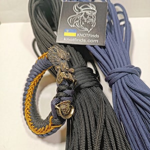 May include: A black and blue paracord bracelet with a gold Viking-style pendant. The bracelet is made with a cobra stitch and has a yellow accent. Two spools of paracord are also shown in the image, one black and one blue.