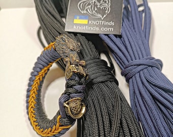 Puede incluir: Un brazalete de paracord negro y azul con un colgante dorado de estilo vikingo. El brazalete está hecho con una puntada de cobra y tiene un acento amarillo. También se muestran dos carretes de paracord en la imagen, uno negro y uno azul.