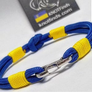 May include: A blue and yellow bracelet with a silver clasp. The bracelet is made of blue rope with yellow accents. The clasp is silver. The bracelet is a close-up shot.