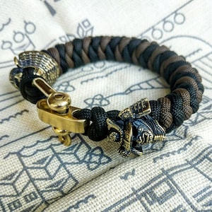 May include: A black and brown paracord bracelet with a brass clasp and a brass samurai helmet charm.