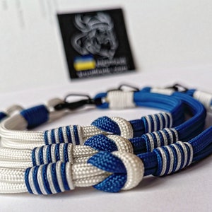 May include: Two braided rope bracelets, one white with blue stripes and one blue with white stripes, are tied together with a knot. The bracelets are made of paracord and have a metal clasp.