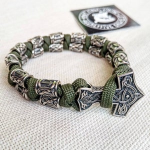 May include: A green paracord bracelet with silver metal beads and a silver Mjolnir charm. The beads have a knot design and the charm has a detailed design of a hammer with a knot design.