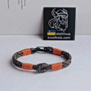 May include: A black and orange paracord bracelet with a black metal clasp. The bracelet has a knot in the center and is made with two different types of paracord.