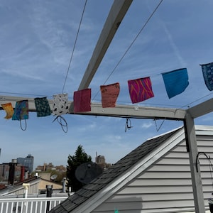 May include: A string of colorful fabric flags hanging from a white wooden pergola. The flags are made of different materials and have various patterns. The flags are hanging against a blue sky with white clouds.