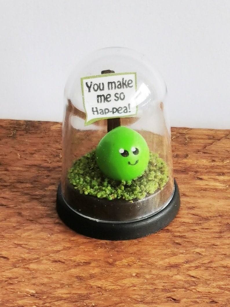 May include: A green pea with a smiley face is inside a glass dome with a sign that reads "You make me so hap-pea!"
