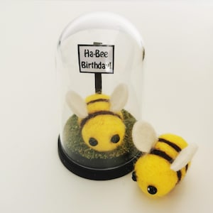 May include: A yellow felt bee with black stripes and white wings, next to a similar bee inside a glass dome. The dome has a sign that reads "Ha-Bee Birthday!" on a green base.