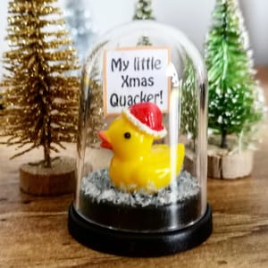May include: A yellow rubber ducky wearing a red Santa hat inside a glass dome. The duck is sitting on a bed of gray material. A sign reads "My little Xmas Quacker!" with Christmas trees in the background.