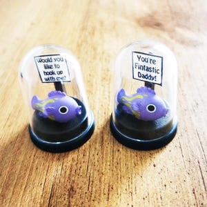 May include: Two glass dome ornaments with purple and yellow fish figurines inside. One dome has a sign that reads "Would you like to hook up with me?" and the other dome has a sign that reads "You're Fintastc Daddy!"