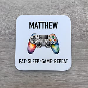 May include: A white square coaster with a vibrant video game controller illustration. The name "MATTHEW" is printed in black above the controller, with the phrase "EAT • SLEEP • GAME • REPEAT" below.