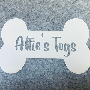 Op de afbeelding: Een wit bord in de vorm van een bot met de tekst "Alfie's Toys" in een cursief schrift.