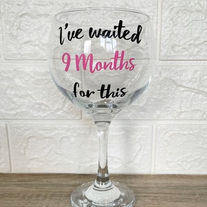 Op de afbeelding: Een helder glas met een steel en een ronde basis. Het glas heeft een zwarte vinyl sticker met de tekst "I've waited 9 Months for this" met het nummer 9 in roze.