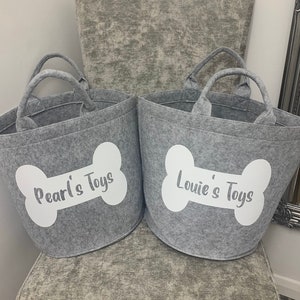 Two grey felt storage bins with white bone-shaped designs and the text "Pearl's Toys" and "Louie's Toys" printed on them.