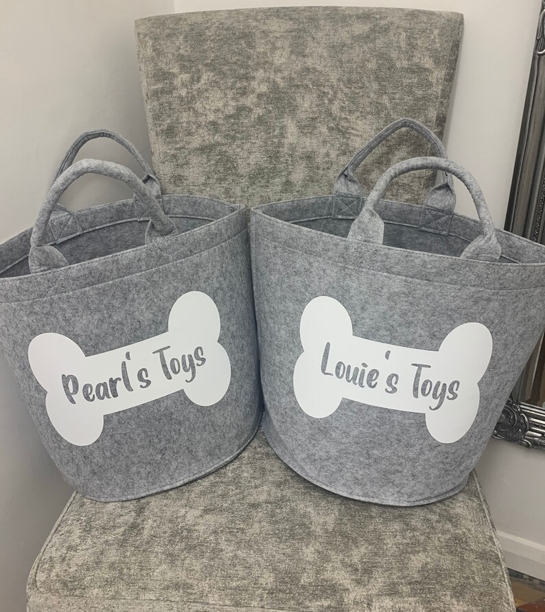 Two grey felt storage bins with white bone-shaped designs and the text "Pearl's Toys" and "Louie's Toys" printed on them.