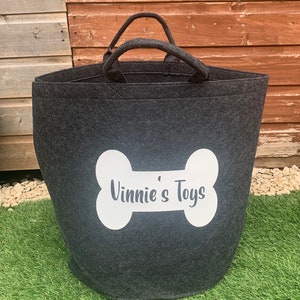 A charcoal grey felt storage bin with a white bone-shaped design that says "Vinnie's Toys". The bag has two handles and is sitting on a green grassy surface.