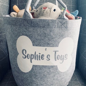 Personalised Dog Toy Storage Basket: Bone Design, Grey Bag With Handles. A grey felt storage bin with a white bone-shaped design that says "Sophie's Toys". The bag has two handles.