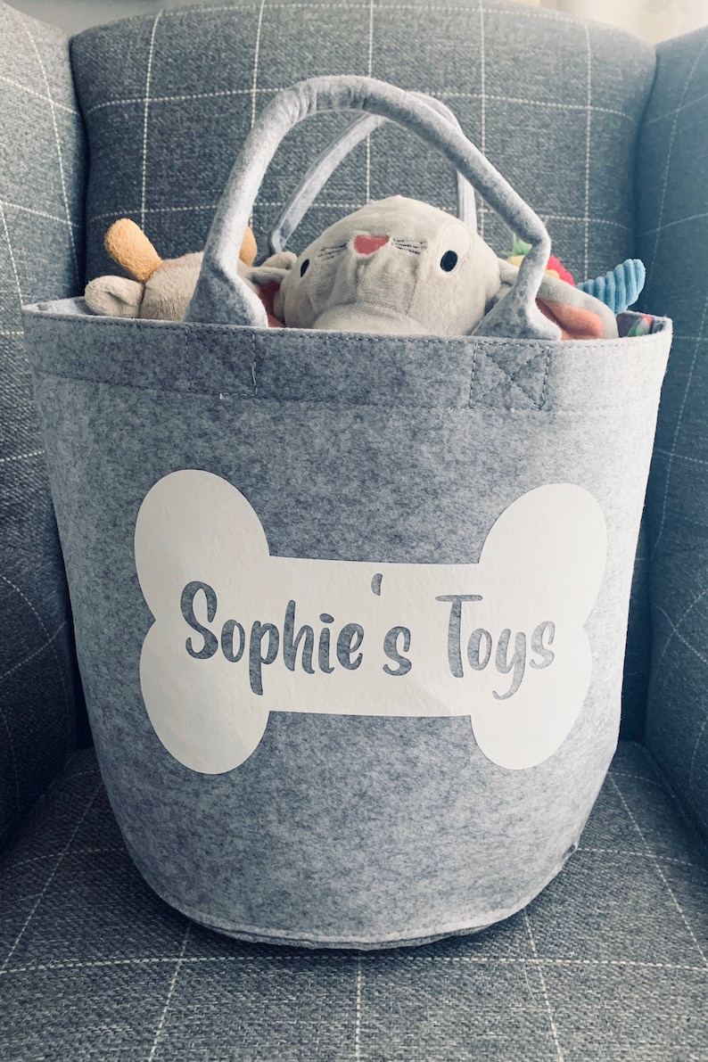 Personalised Dog Toy Storage Basket: Bone Design, Grey Bag With Handles. A grey felt storage bin with a white bone-shaped design that says "Sophie's Toys". The bag has two handles.