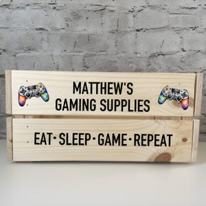 May include: A wooden crate with the text "MATTHEW'S GAMING SUPPLIES" and "EAT SLEEP GAME REPEAT" on it. The crate is decorated with two colorful video game controllers.