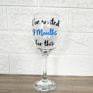 Op de afbeelding: Een helder glazen wijnglas met een zwarte en blauwe vinyl sticker met de tekst "Ik heb 9 maanden gewacht op dit moment".