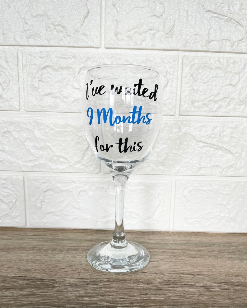 Op de afbeelding: Een helder glazen wijnglas met een zwarte en blauwe vinyl sticker met de tekst "Ik heb 9 maanden gewacht op dit moment".