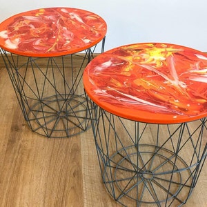 May include: Two round side tables with a wire basket base and a vibrant orange top. The table tops feature a swirling pattern of red, orange, yellow, and white. The wire bases are dark gray.
