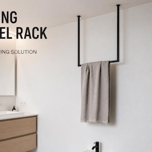 Può includere: Un portasciugamani da soffitto nero con un asciugamano beige appeso. Il testo "CEILING TOWEL RACK" e "SPACE SAVING SOLUTION" sono visualizzati in nero. Il bagno ha una parete bianca, un lavandino bianco e un mobile in legno.