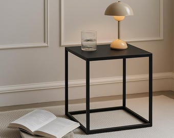 May include: A black metal side table with a square top and a glass of water and a beige table lamp with a mushroom-shaped shade on top. The table is on a white rug with a subtle texture.