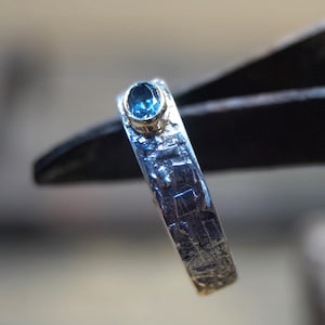 May include: A silver ring with a textured band and a small, oval, blue gemstone. The ring is held by a black tool, with a blurred background. The ring is a piece of jewelry.