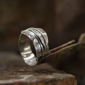 Könnte beinhalten: Ein silberner Ring mit einem strukturierten Federdesign. Der Ring wird von einer Zange gehalten.