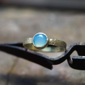 Peut inclure: Une bague en or avec une pierre précieuse ronde bleu clair. La bague est sertie sur un anneau texturé et est maintenue par une pince en métal noir. La bague est un bijou.