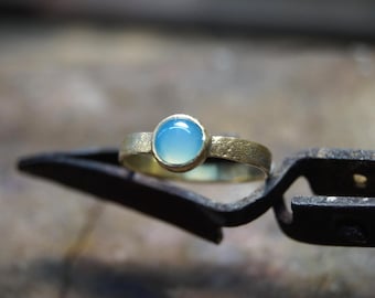 Può includere: Un anello d'oro con una gemma rotonda blu chiaro. L'anello è incastonato su una fascia strutturata ed è tenuto da un paio di pinze in metallo nero. L'anello è un gioiello.