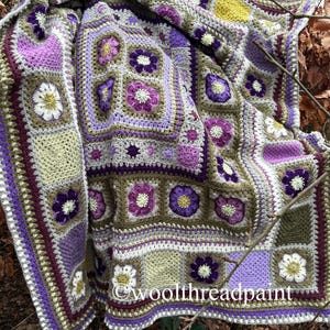 May include: A handmade crochet blanket in shades of purple, white, green, and brown. The blanket features a floral pattern with square and rectangular motifs. The text "woolthreadpaint" is visible on the blanket.