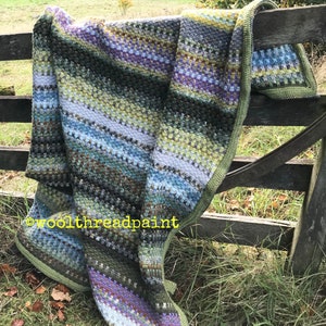 Könnte beinhalten: Eine mehrfarbige Häkeldecke mit Streifen in Grün, Blau, Lila und Weiß. Die Decke ist über einen Holzzaun gelegt.