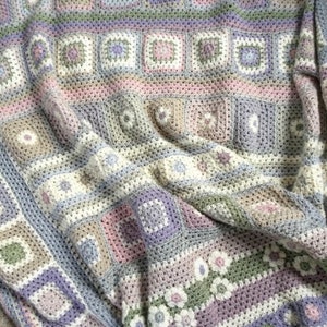 Pode incluir: Uma manta de crochê com um padrão texturizado em tons de rosa, roxo e verde. A manta é feita com uma variedade de pontos e tem uma sensação macia e aconchegante.
