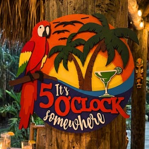 May include: A circular wooden wall sign featuring a red parrot perched on a branch, palm trees, and a cocktail glass against a sunset background. The text reads It's 5 O'CLOCK somewhere in red and white lettering, layered over a blue wave design.