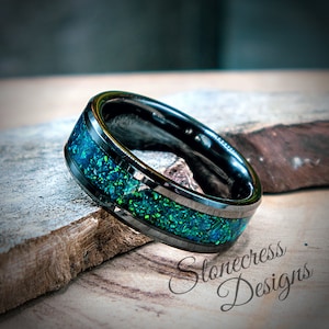 May include: A black tungsten carbide ring with a green and blue opal inlay and a polished finish. The ring has a beveled edge and a flat top. The ring is on a wooden surface. Stonecress Designs.