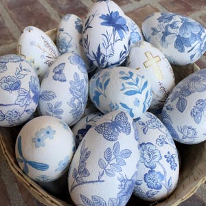 May include: A collection of white decorative eggs with blue floral and butterfly patterns. Some eggs feature a gold cross design. The eggs are arranged in a woven basket, creating a charming display.