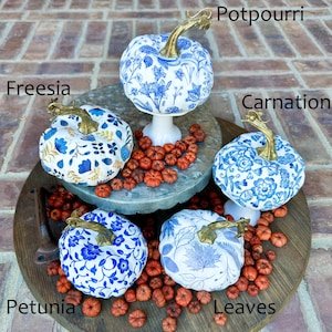Puede incluir: Cinco calabazas blancas decoradas con motivos florales azules y dorados. Las calabazas est&aacute;n dispuestas en un soporte de madera de dos niveles con potpourri rojo esparcido a su alrededor. Las calabazas est&aacute;n etiquetadas con los nombres de las flores: Freesia, Clavel, Potpourri, Petunia y Hojas.