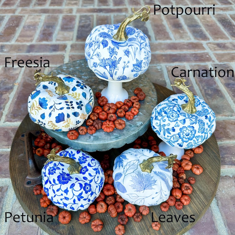 Puede incluir: Cinco calabazas blancas decoradas con motivos florales azules y dorados. Las calabazas est&aacute;n dispuestas en un soporte de madera de dos niveles con potpourri rojo esparcido a su alrededor. Las calabazas est&aacute;n etiquetadas con los nombres de las flores: Freesia, Clavel, Potpourri, Petunia y Hojas.