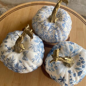 Peut inclure: Trois citrouilles d&eacute;coratives avec des motifs floraux blancs et bleus et des tiges dor&eacute;es. Les citrouilles sont dispos&eacute;es sur une surface en bois, mettant en valeur un th&egrave;me d'automne ou de r&eacute;colte. Les motifs bleus varient.