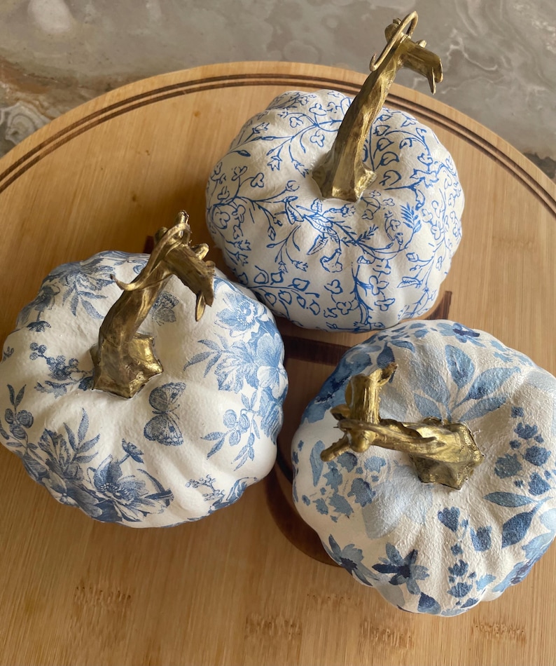 Peut inclure: Trois citrouilles d&eacute;coratives avec des motifs floraux blancs et bleus et des tiges dor&eacute;es. Les citrouilles sont dispos&eacute;es sur une surface en bois, mettant en valeur un th&egrave;me d'automne ou de r&eacute;colte. Les motifs bleus varient.